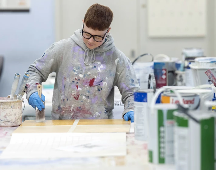 Focused student in paint-splattered hoodie painting on a wooden board Focused student in paint-splattered hoodie painting on a wooden board